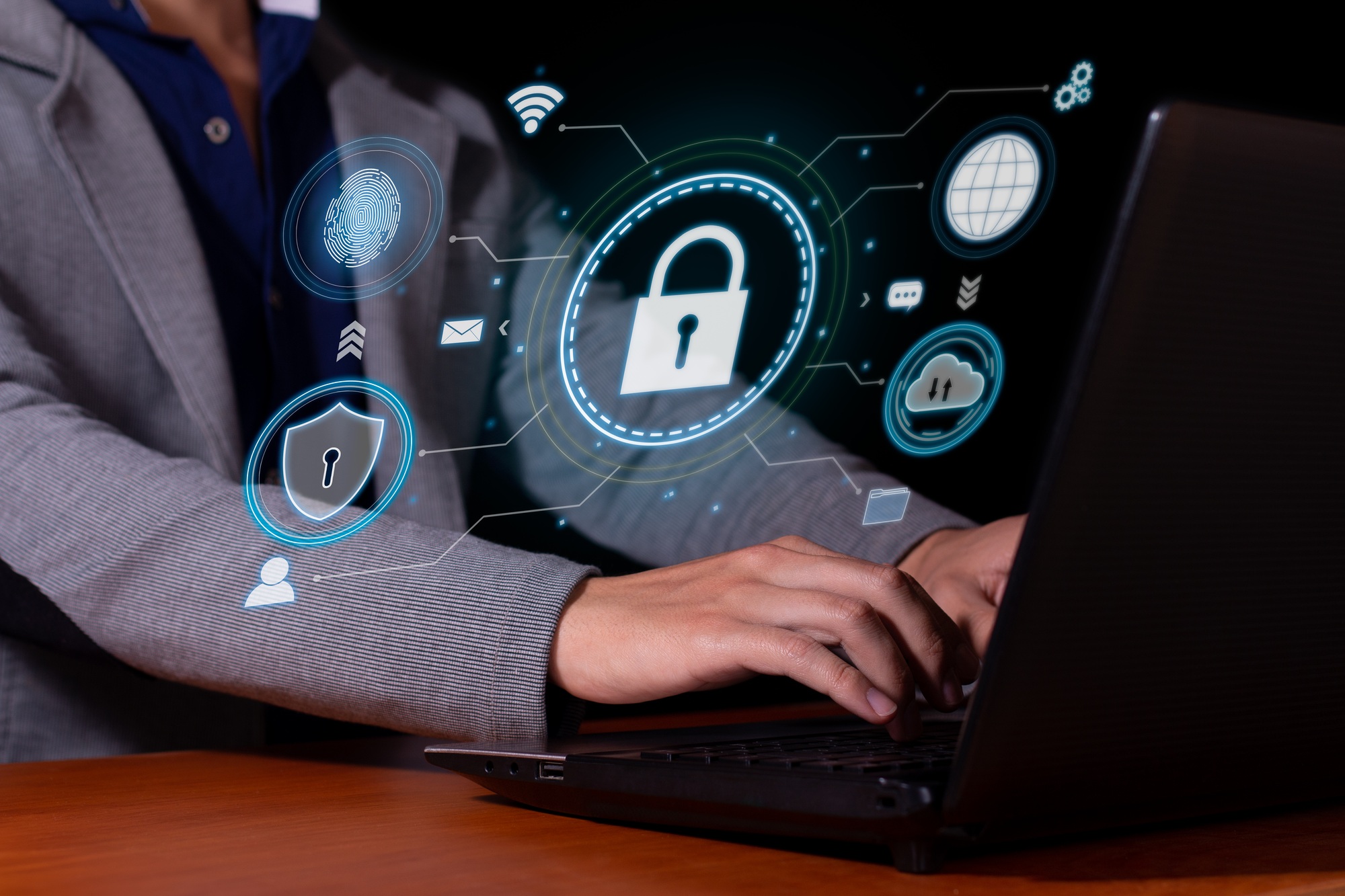 Hands typing on a laptop keyboard with floating digital security icons (padlock, shield, fingerprint, cloud and user).