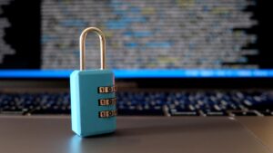 A small blue combination padlock sitting on a laptop keyboard with blurred code visible on the screen behind it.